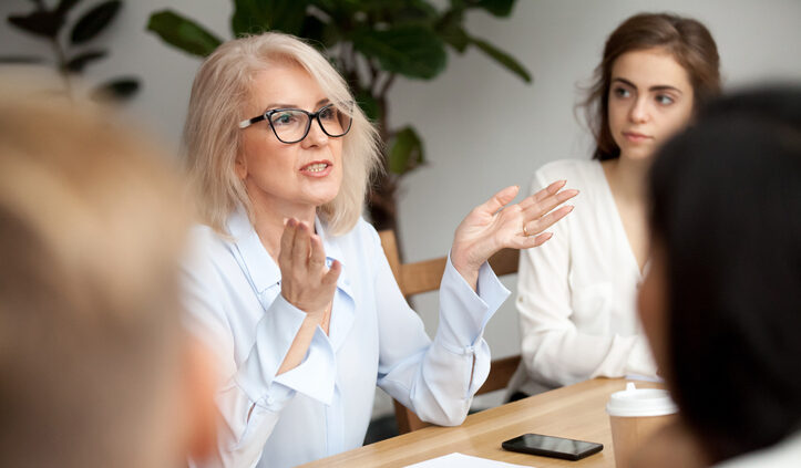 Todays leaders need to embrace intelligent enterprise search. A blond woman in her fifties with black rimmed glasses leads a team meeting around a conference table.