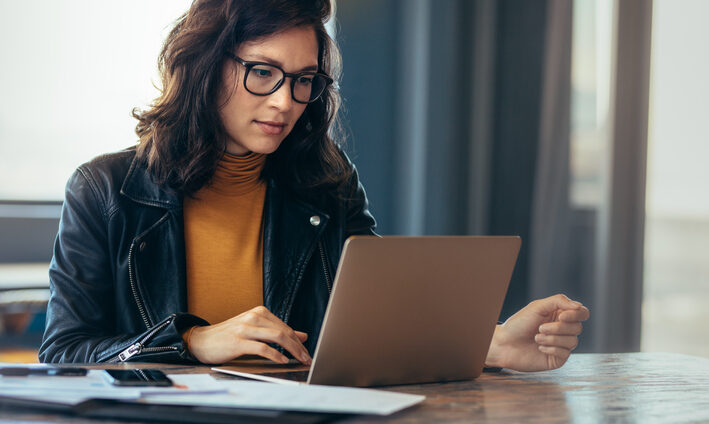 A young woman with brown hair and glasses works on her laptop.