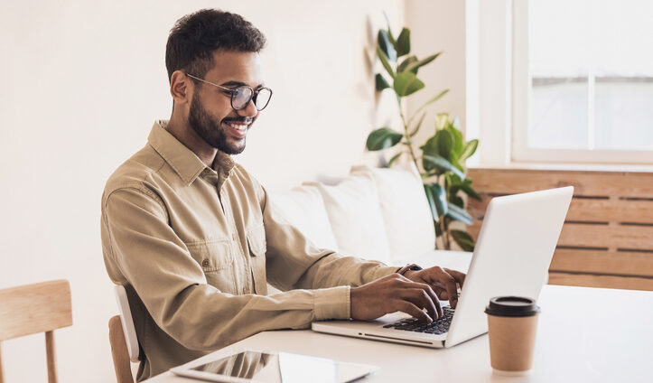 Young man in glasses happily works at his computer in a warmly daylit room.