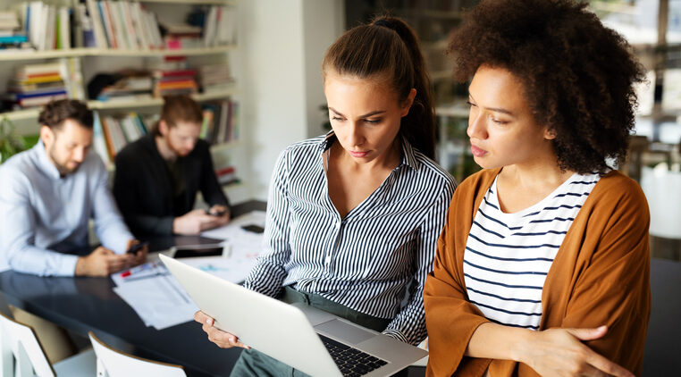 Two women colleagues look at a laptop screen. Two men work in the background.