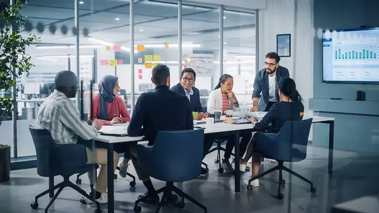 A group of office workers are seated at a round meeting table where they discuss their knowledge management.