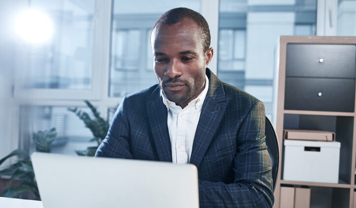 Smart businessman. Portrait of serious young elegant Black man in a suit is sitting at table in modern office and working on laptop. He is looking at the screen with concentration. Strategic research portal.