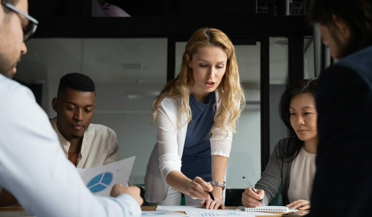 Concentrated young businesswoman explaining market research results in graphs to mixed race colleagues. Focused group of diverse employees holding brainstorming meeting, discussing project ideas. Competitive intelligence.