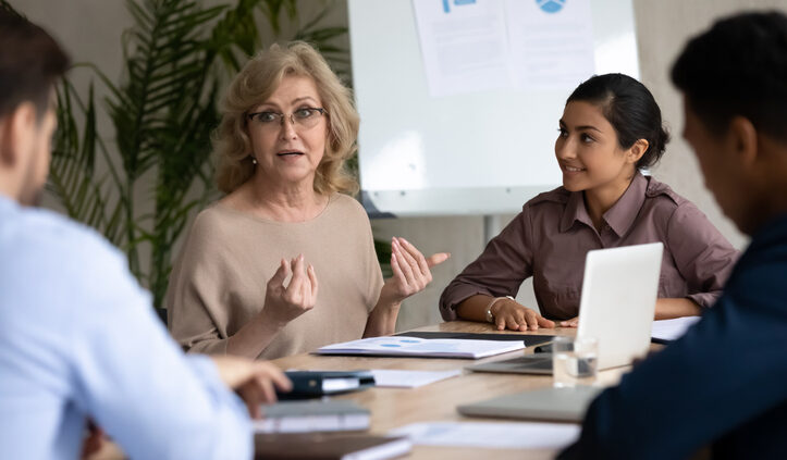 Mature 60 year old businesswoman with indian ethnic partner talking in boardroom at meeting. Confidence woman training diverse corporate team at briefing. Young employee share thoughts sitting. Knowledge management systems