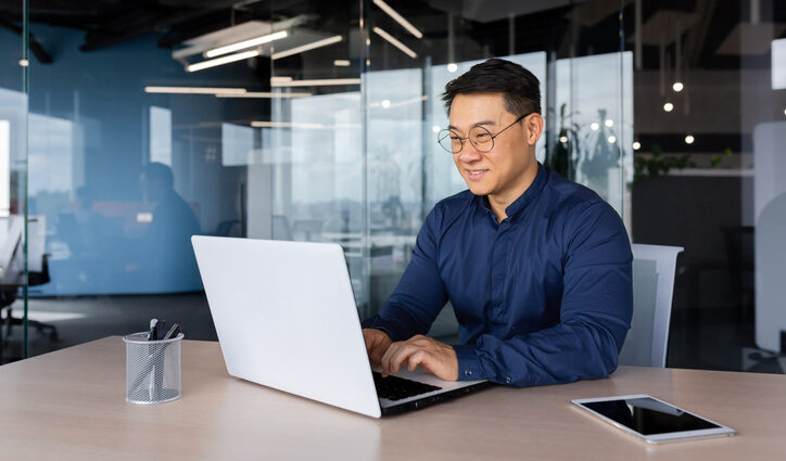 Successful mature asian working inside office using laptop, man typing on keyboard and smiling, businessman in shirt and glasses satisfied with work and achievement results, programmer at work. Generative AI and copyright.