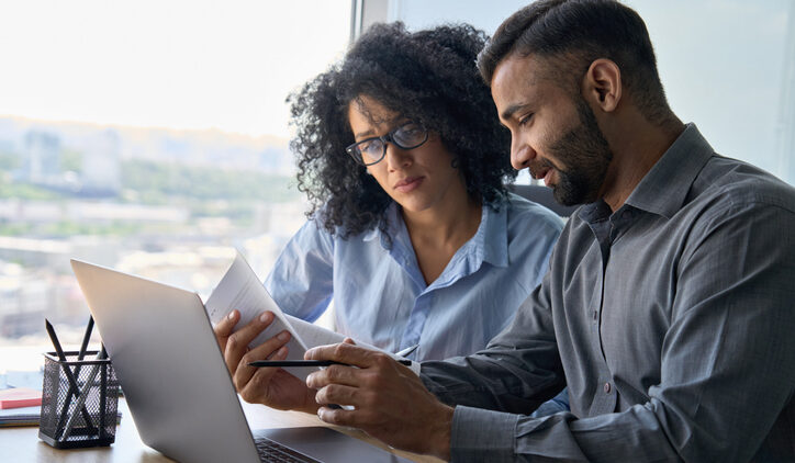 Multiethnic male indian mentor and female African American intern sitting at desk with laptop doing paperwork together discussing project financial report. Corporate business collaboration concept. For competitive intelligence research, curated content and curated sources matter.